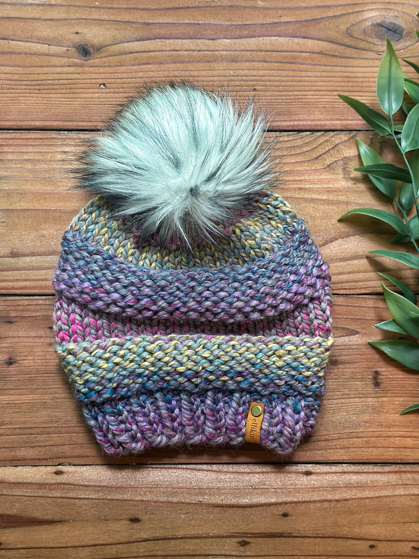 A multicolor striped knit beanie with a large mint green faux fur pom-pom, laid flat on a rustic wooden surface next to green leaves.