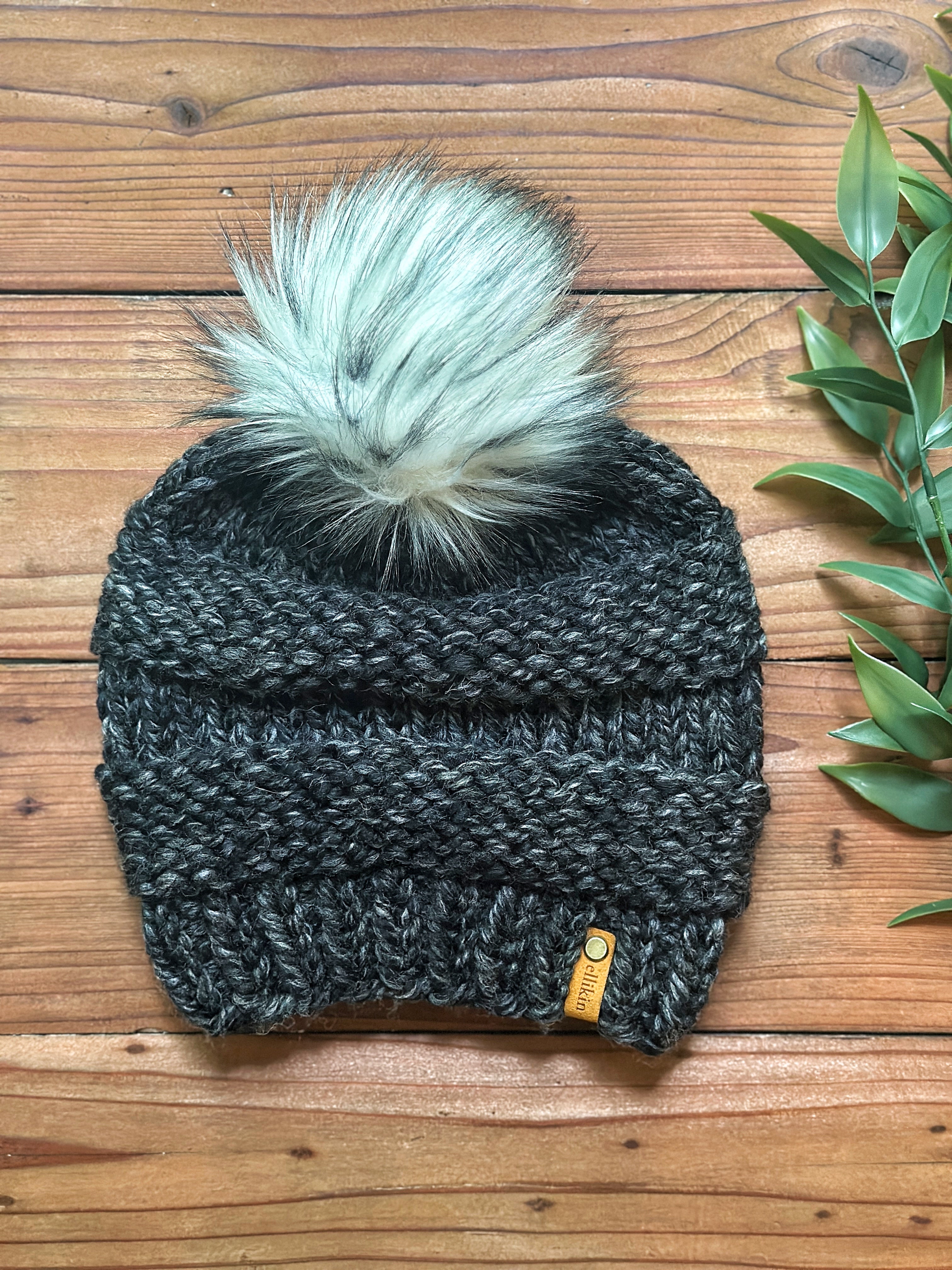 A dark charcoal gray knit beanie with a large, fluffy pale sage green and white pom-pom, laid flat on a rustic wooden background next to green leaves.