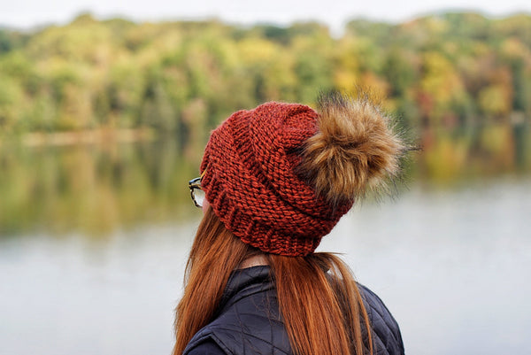 A person viewed from behind wearing a chunky rust-red knit beanie with a brown faux-fur pom-pom, standing by a calm lake with a blurred autumn forest in the background.