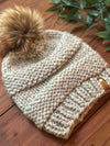 A chunky knit beanie hat in a warm oatmeal beige color, featuring a large brown faux-fur pom-pom on top and a small tan leather brand tab on the side, displayed on a wooden surface with green leaves in the background.