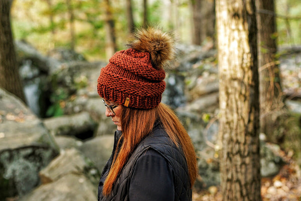 A woman wearing a rust-brown chunky knit beanie with a brown faux-fur pom-pom, standing outdoors in a wooded area with rocks and trees in the background.