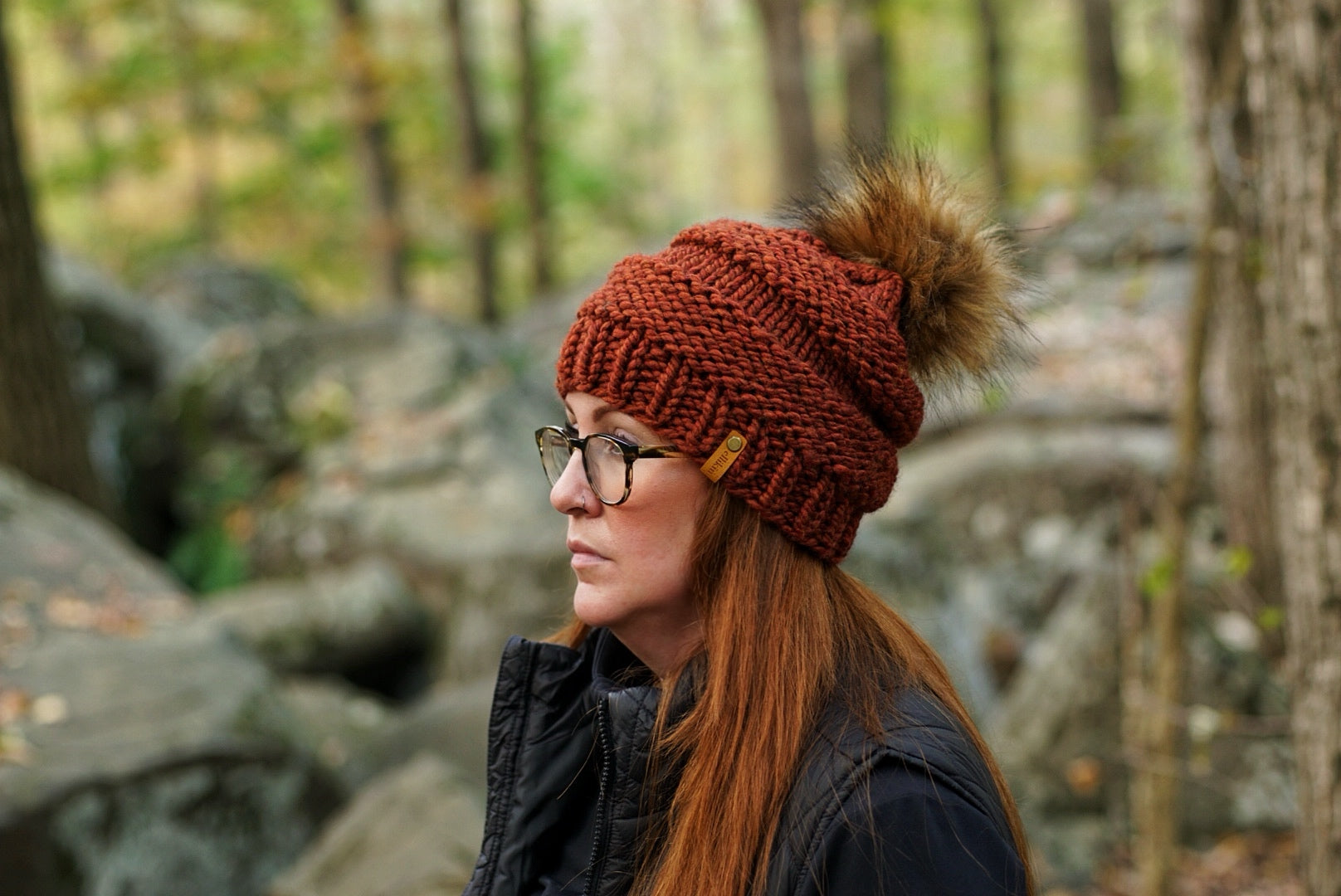 A woman in profile wearing a chunky knit brown beanie with a brown faux-fur pom-pom and a small gold tag, photographed outdoors in a blurred forest setting.