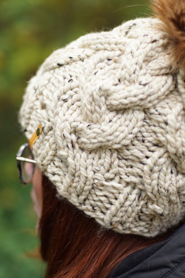 Close-up of a person wearing a chunky cream cable-knit beanie with a brown faux-fur pom-pom, photographed outdoors against a blurred green background.