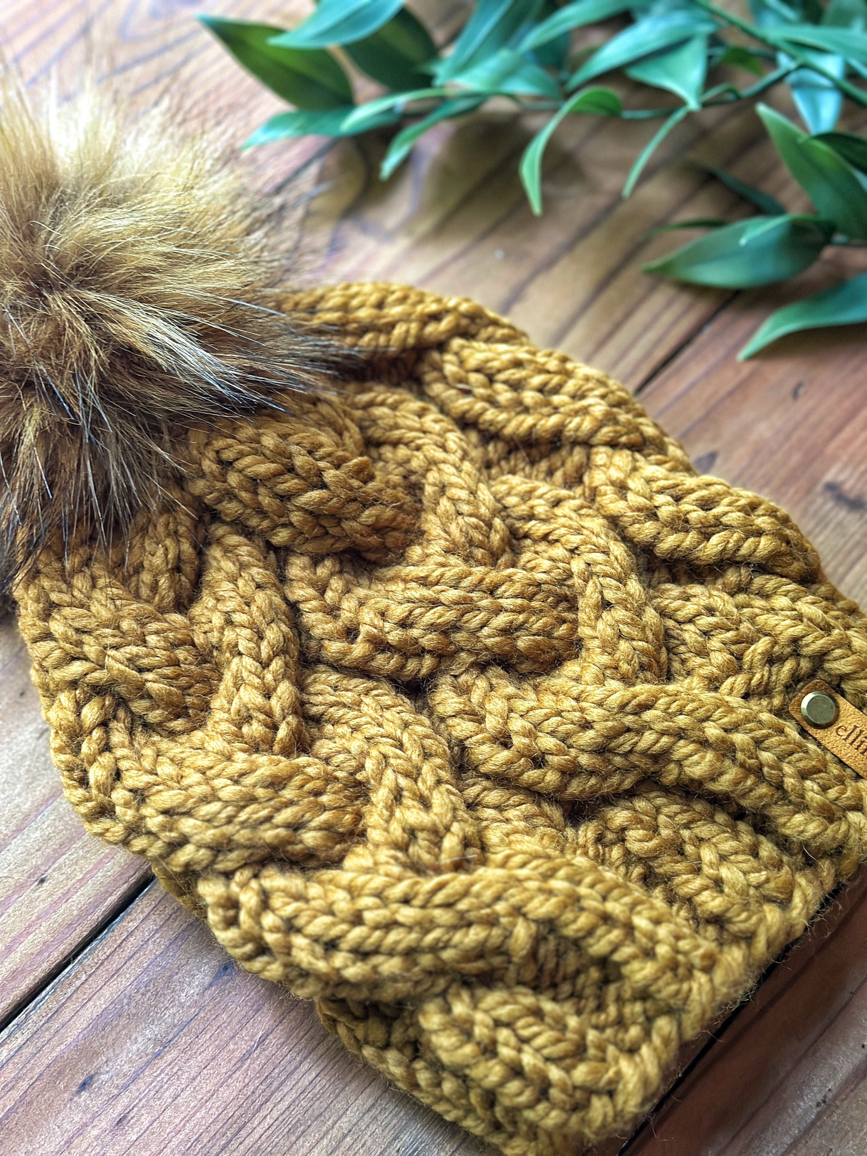 A chunky cable-knit beanie in a warm mustard yellow color, featuring a large brown faux fur pom-pom and a small wooden brand tag, displayed on a rustic wooden surface with green leaves in the background.