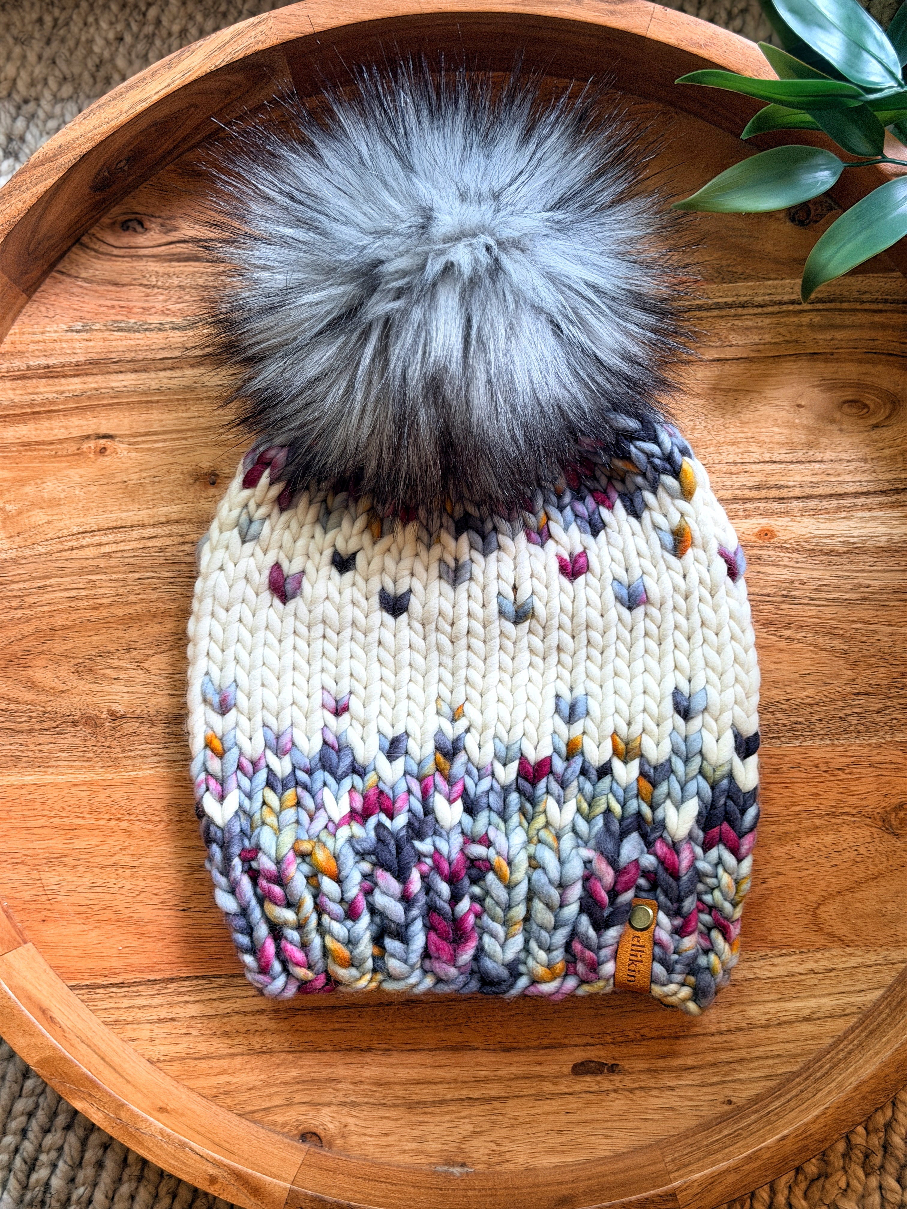 A chunky knit beanie hat with a large fluffy gray pom-pom, featuring a multicolor heart pattern on a cream base, displayed on a round wooden tray.