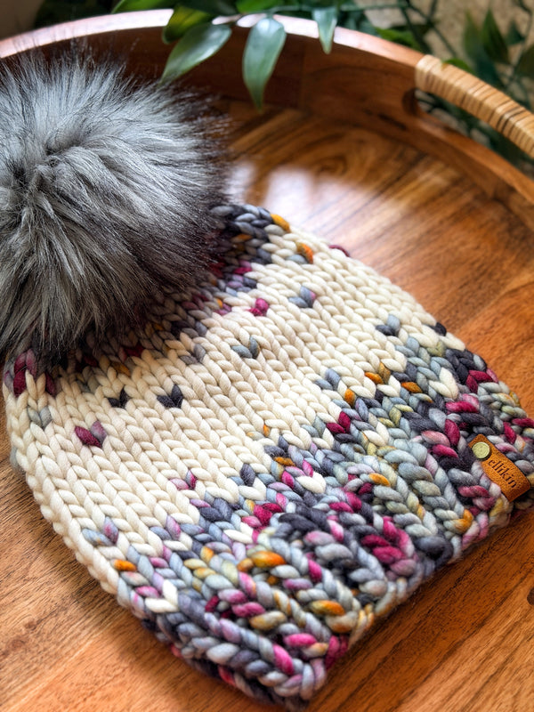 A chunky knit beanie hat with a large gray faux fur pom-pom, resting on a round wooden tray. The beanie features a cream base with a variegated pattern of gray, pink, orange, and purple yarns concentrated in the lower half.