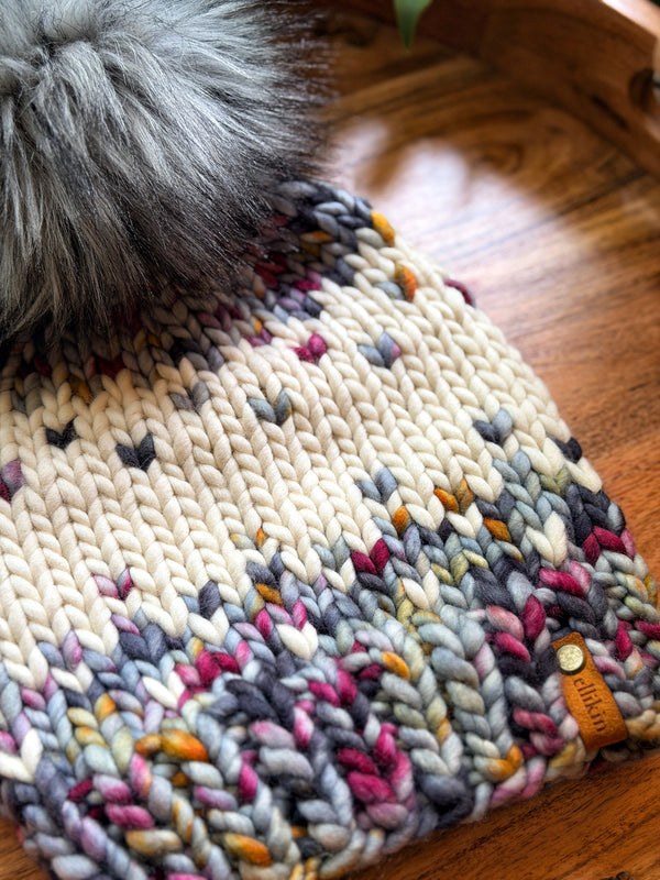 A close-up of a chunky knit beanie with a large gray faux-fur pom-pom, resting on a wooden tray. The beanie features a cream base with variegated yarn sections in gray, magenta, orange, and dark gray, finished with a small tan leather tag.