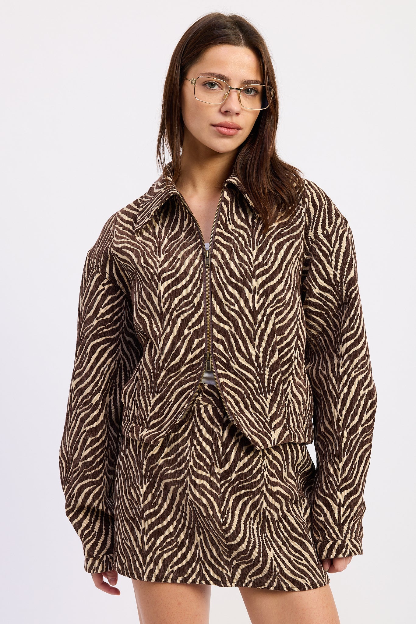 A woman wearing a brown and beige zebra-print jacket with a front zipper, photographed against a plain light gray studio background.