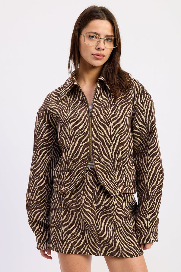 A woman wearing a brown and beige zebra-print jacket with a front zipper, photographed against a plain light gray studio background.