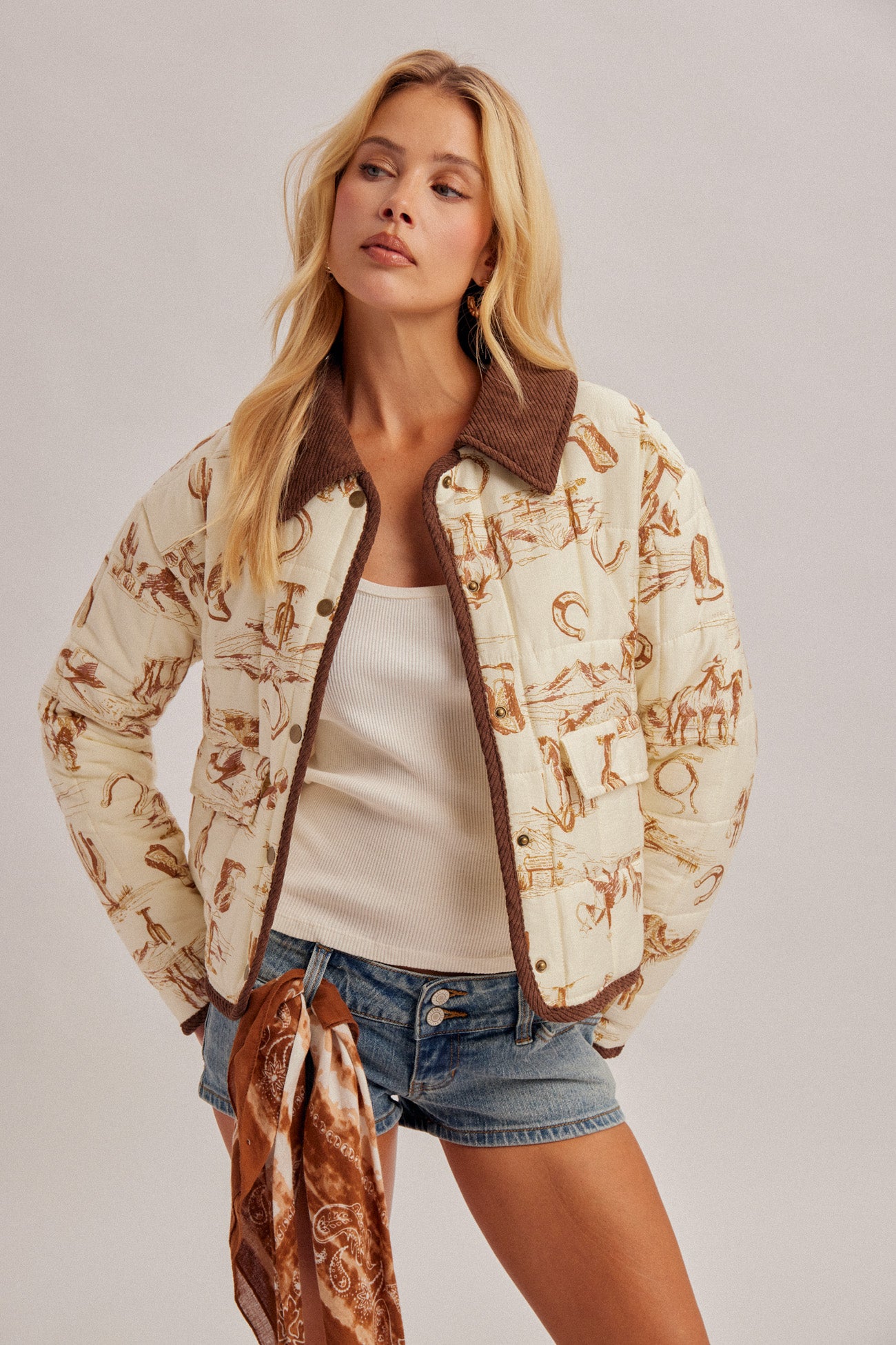A woman models a cream-colored quilted bomber jacket with a brown patterned print, brown suede-look collar and trim, worn over a white tank top and denim shorts.