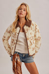 A woman models a cream-colored quilted bomber jacket with a brown patterned print, brown suede-look collar and trim, worn over a white tank top and denim shorts.