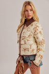A blonde model wearing a cream-colored quilted jacket with a brown western-themed print and a brown faux-shearling collar, styled with denim shorts and a matching brown scarf against a light gray studio background.