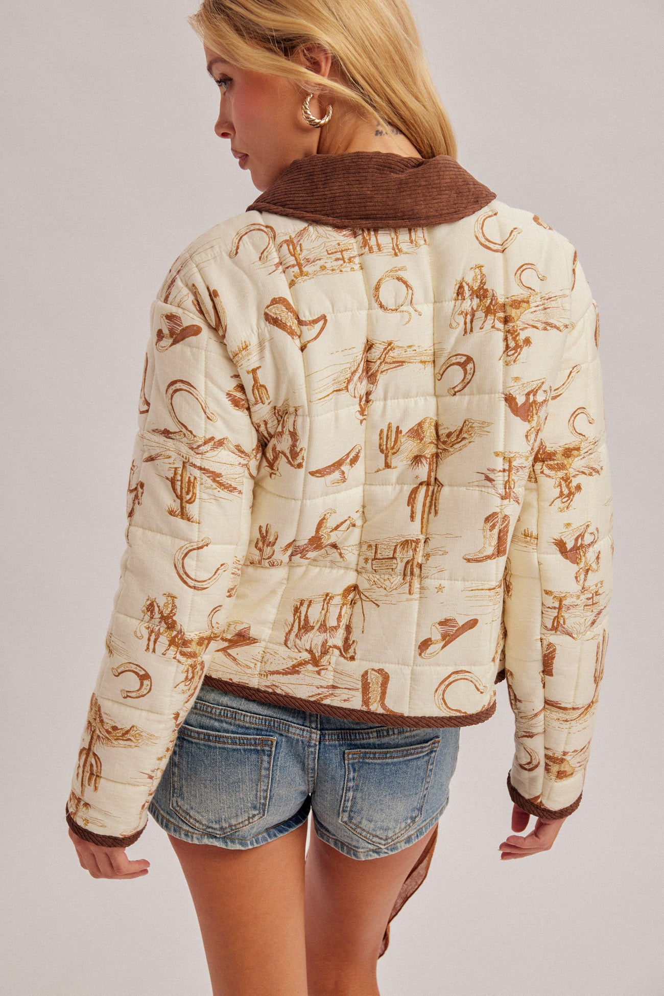 A woman shown from the back wearing a cream quilted jacket with a brown corduroy collar and a brown western-themed print, paired with denim shorts, against a light gray studio background.