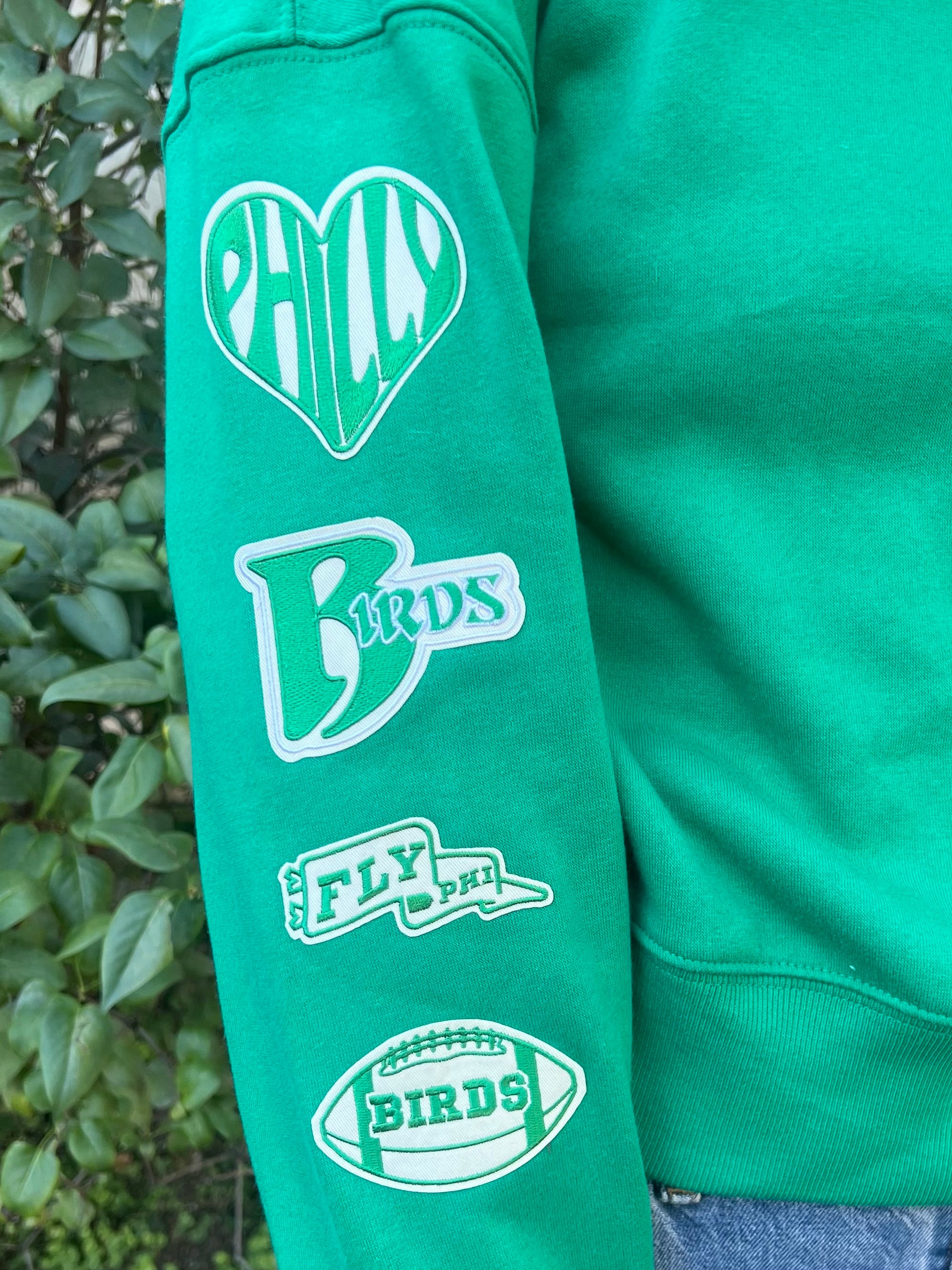 A green sweatshirt sleeve featuring green and cream patches. One is a heart with philly written inside, the next is birds with a large B. The next patch is a waving pennant that says fly phi and the last patch is a football that says birds
