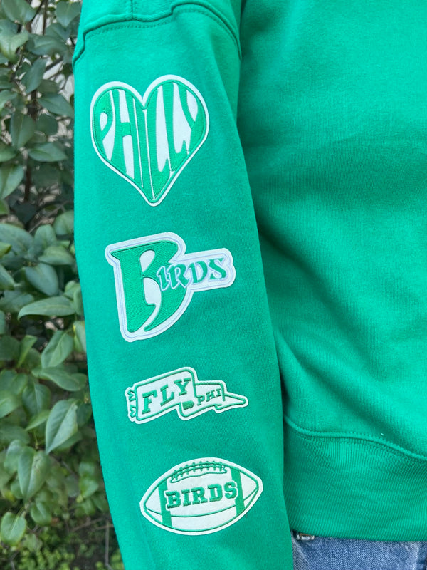 A green sweatshirt sleeve featuring green and cream patches. One is a heart with philly written inside, the next is birds with a large B. The next patch is a waving pennant that says fly phi and the last patch is a football that says birds