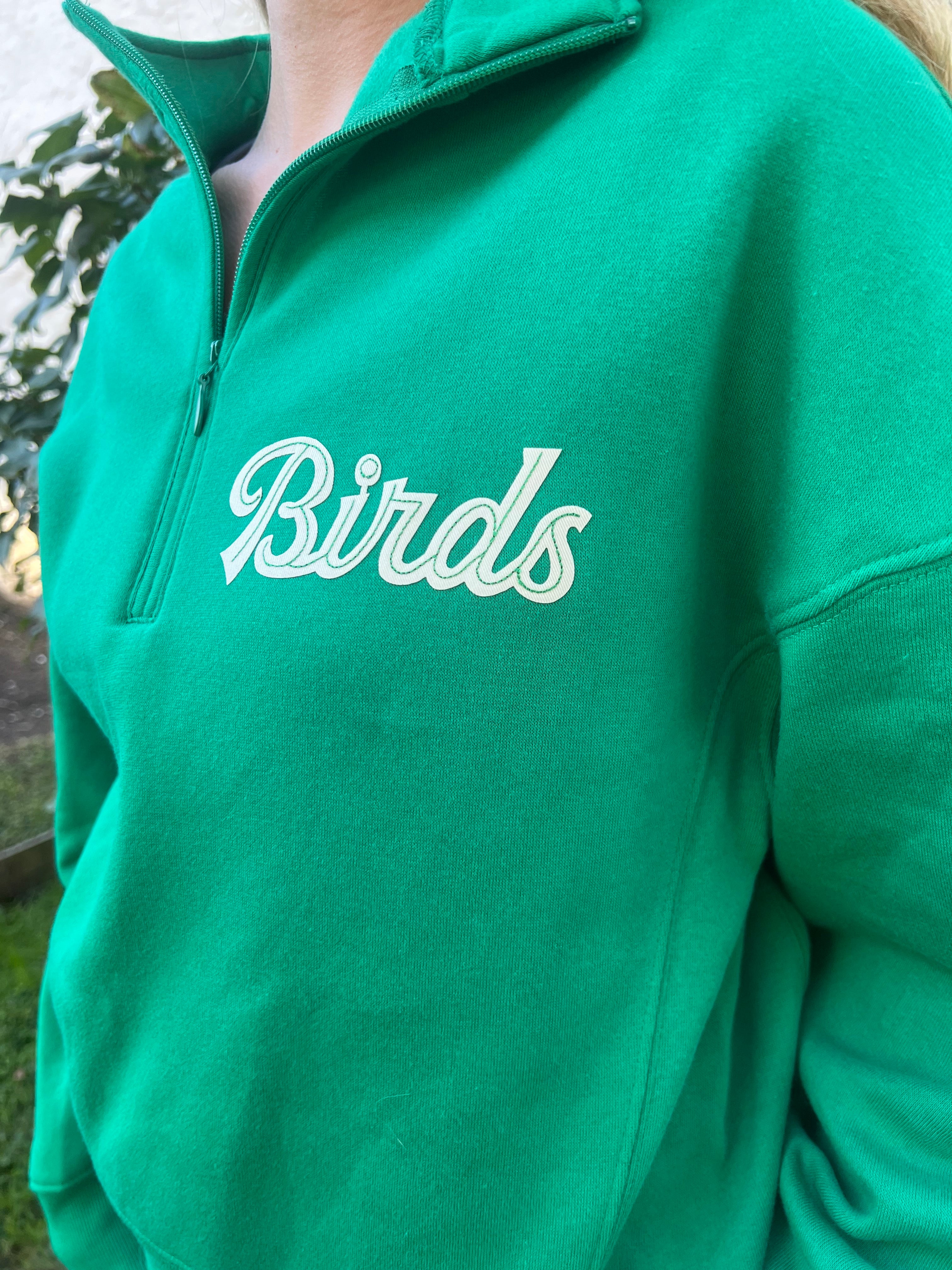 Zoom detail on a green hoodie showing the birds detail in cream lettering and subtle embroidery