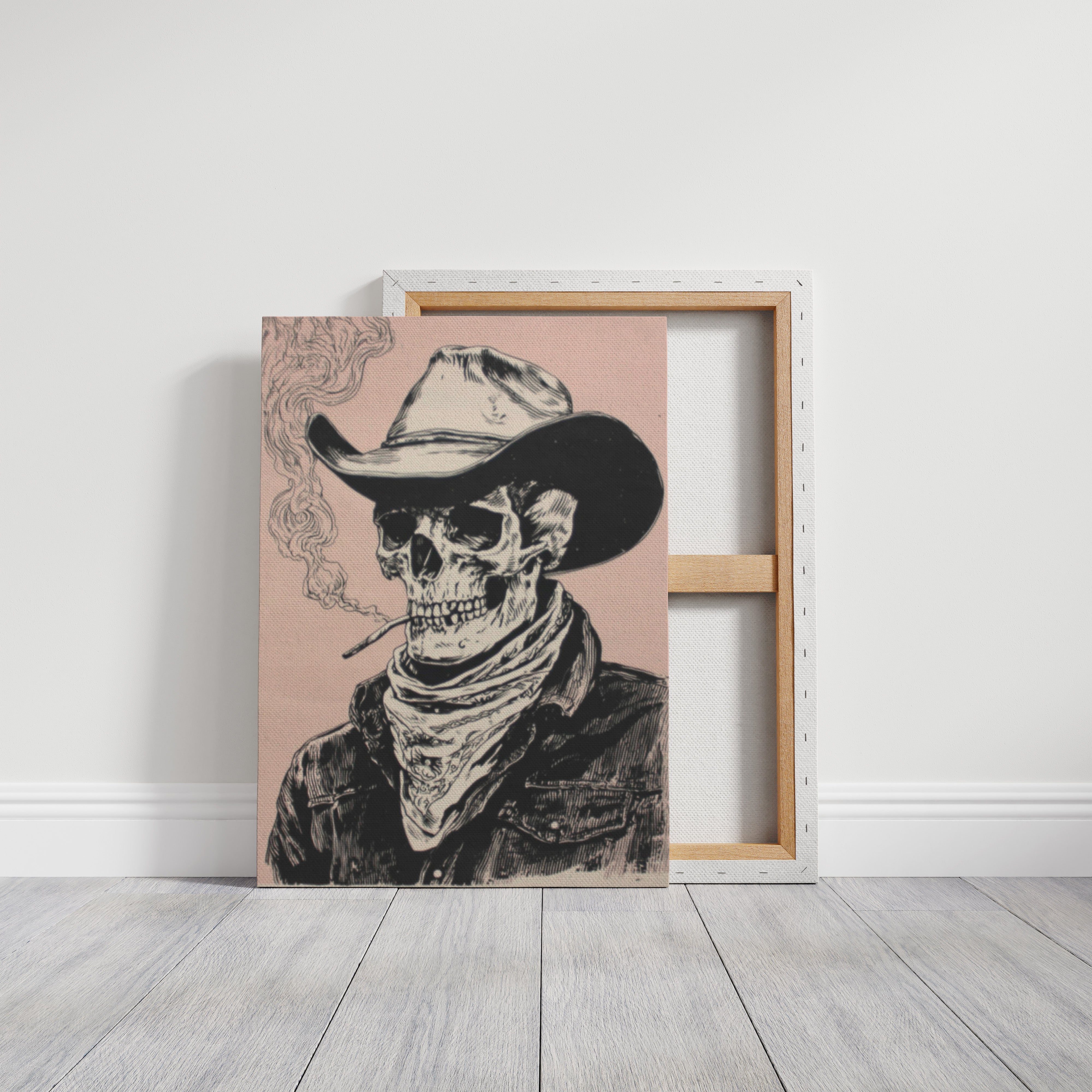 A square canvas print leaning against a white wall on a light wood floor, showing a black ink-style illustration of a cowboy skeleton wearing a hat and bandana. A second empty canvas is visible behind it.