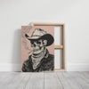 A square canvas print leaning against a white wall on a light wood floor, showing a black ink-style illustration of a cowboy skeleton wearing a hat and bandana. A second empty canvas is visible behind it.
