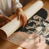 A person's hands and arms are visible as they unroll a large sheet of paper or poster showing a black and white graphic of a skull smoking a cigarette, laid out on a wooden table.