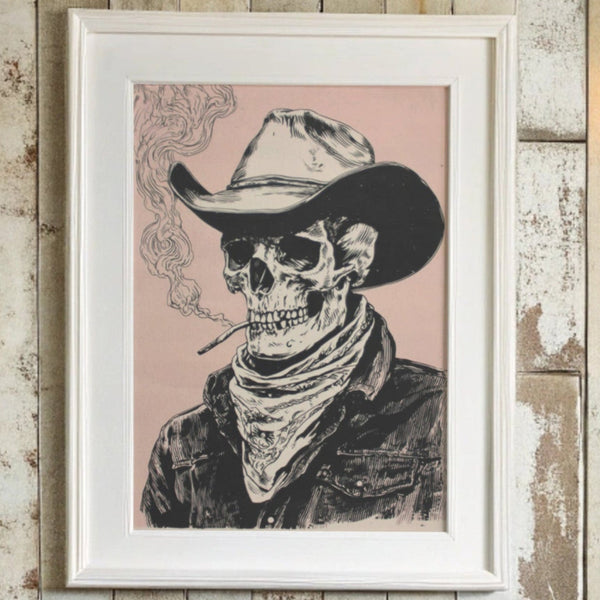 A framed art print featuring a western-style illustration of a skull wearing a cowboy hat and bandana, smoking a cigarette against a dusty pink background, mounted on a rustic whitewashed wooden wall.