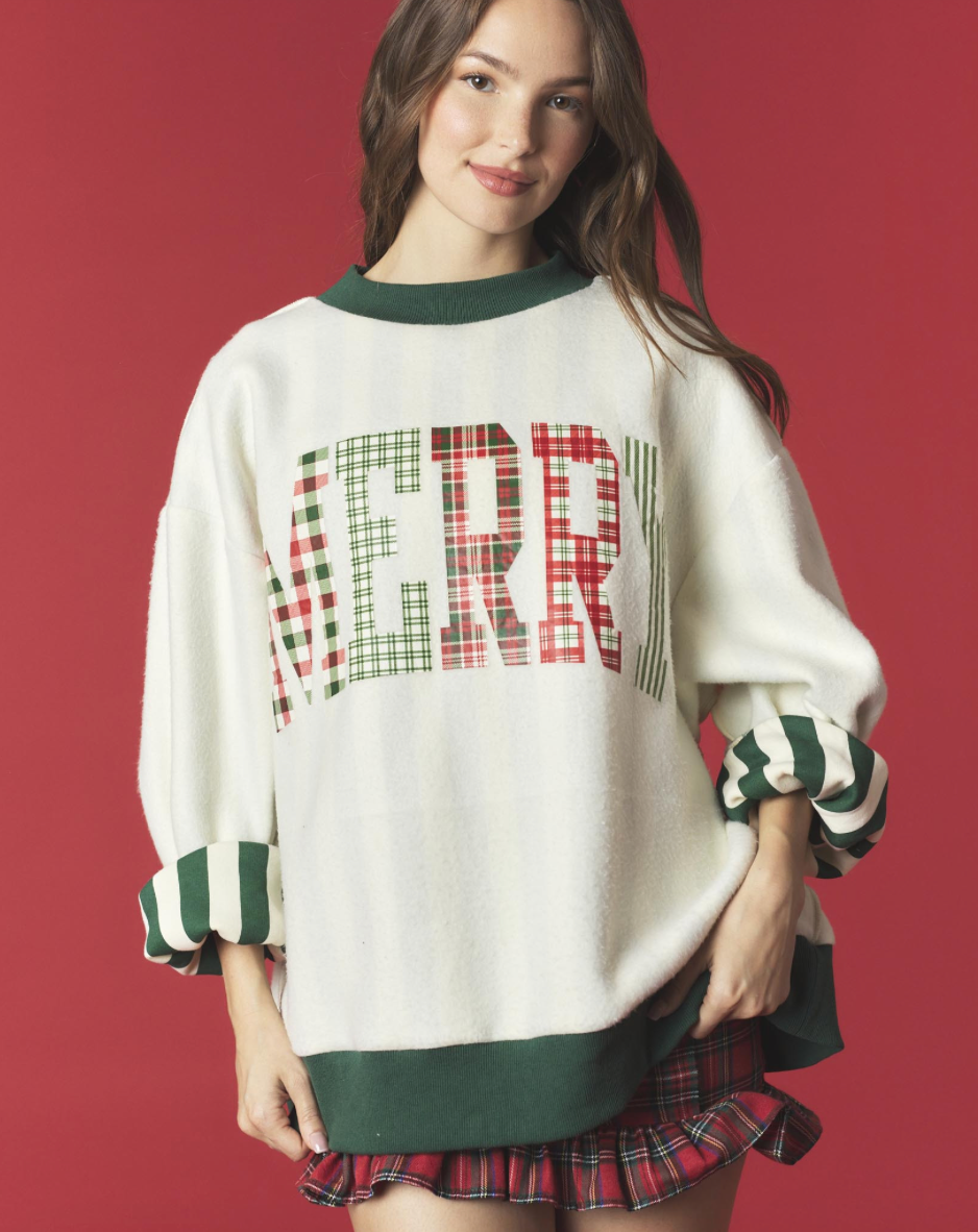 A woman models a holiday-themed cream knit sweater with a green crew neck, green striped cuffs, and a large 'MERRY' graphic across the chest in a red and green plaid pattern. She is also wearing a matching red and green plaid pleated skirt against a solid red studio background.