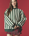 A woman wearing an oversized green and cream vertically striped sweatshirt with a red and green plaid ruffled mini skirt, posed against a solid dark red background.