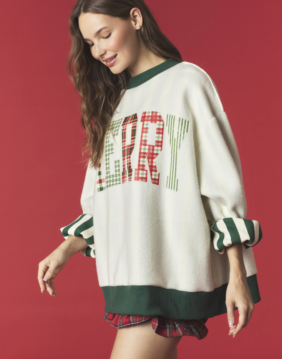 A woman wearing an oversized cream knit sweater with dark green trim and a festive red and green plaid 'MERRY' graphic on the chest, posed against a solid dark red background.