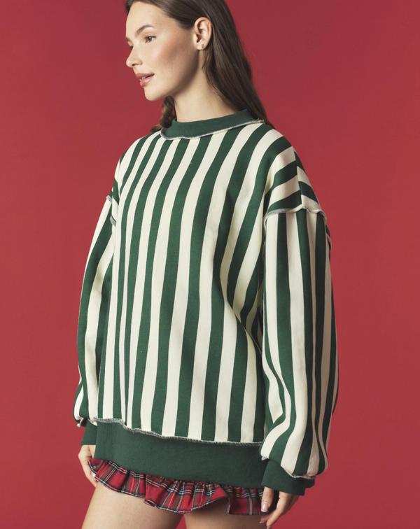 A woman wearing an oversized long-sleeve crewneck sweatshirt with vertical dark green and cream stripes, paired with a red plaid mini skirt, set against a solid dark red studio background.