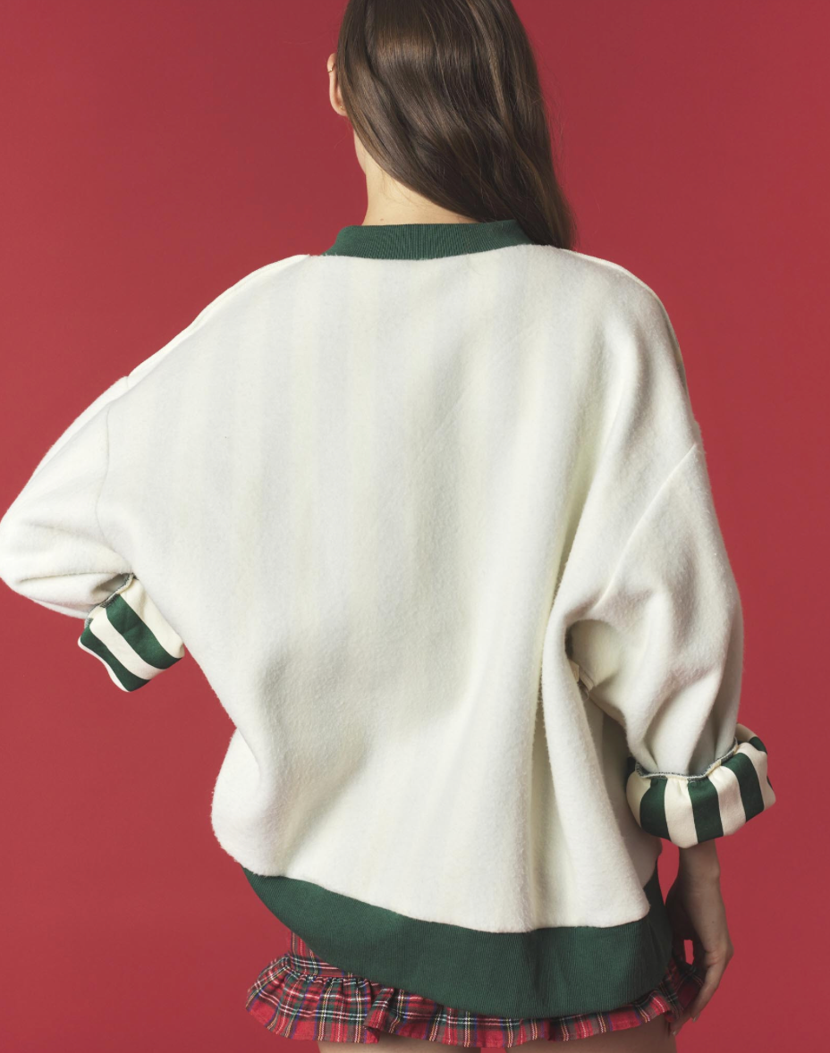 A person shown from the back wearing an oversized cream knit sweater with dark green ribbed trim at the collar, hem, and striped cuffs, against a solid red background.