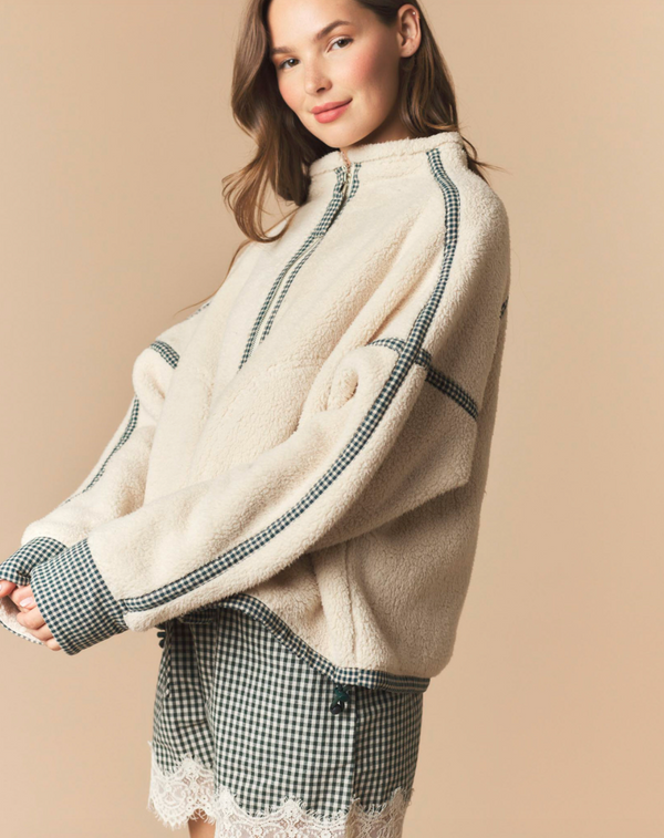 A woman wearing a cream-colored fuzzy quarter-zip pullover sweater with dark green and white gingham patterned stripes, paired with a matching gingham mini skirt, set against a solid beige background.
