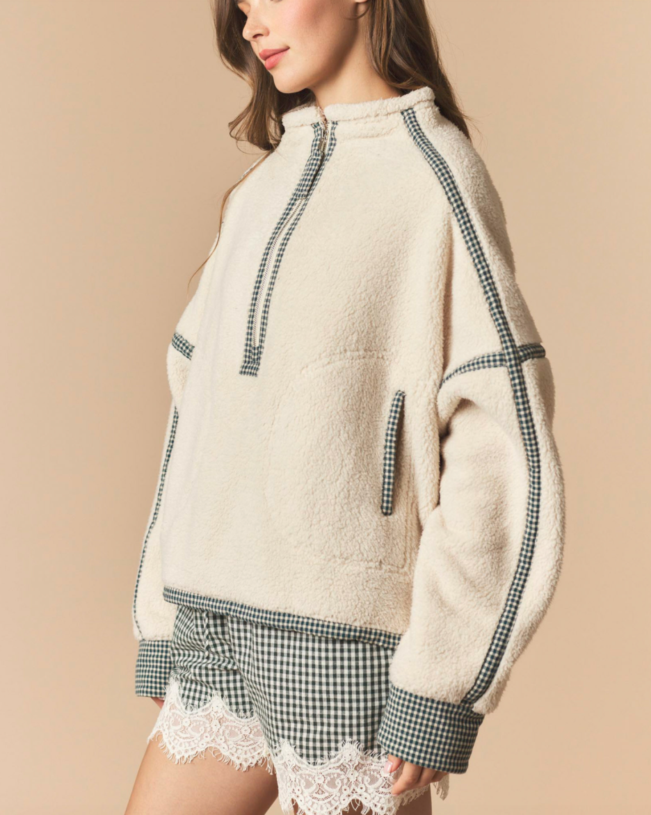 A woman wearing a cream sherpa pullover with black-and-white gingham trim and matching gingham shorts with lace hem, posed against a solid beige studio background.