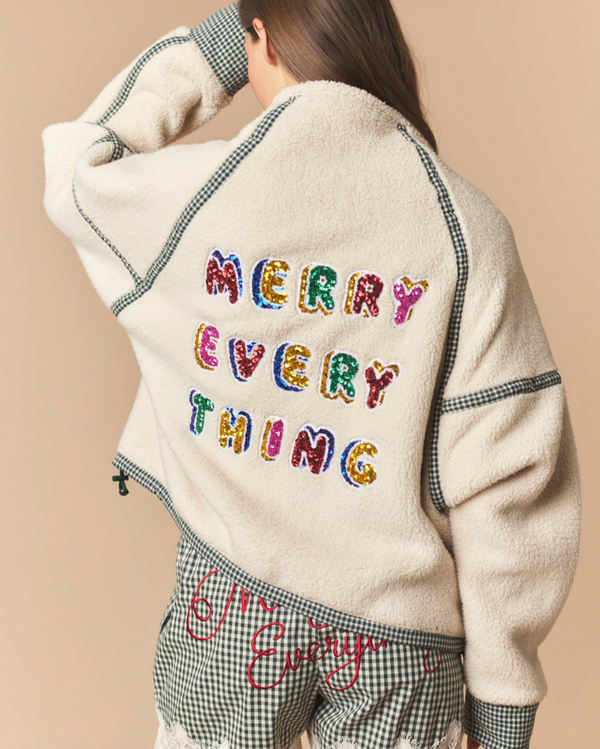 A person shown from the back wearing a cream sherpa jacket with colorful sequined 'MERRY EVERY THING' lettering on the back and a gingham hem, paired with matching gingham shorts against a solid beige background.