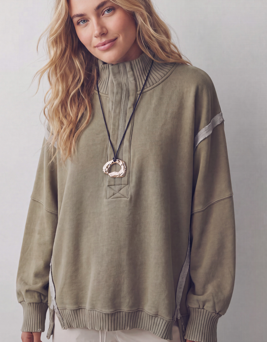 A woman wearing a sage green mock-neck sweatshirt with ribbed cuffs and hem, featuring a decorative geometric embroidery on the chest and a large statement ring necklace.