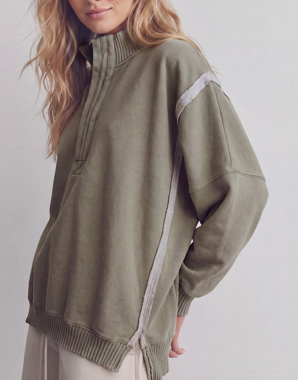 A woman wearing an oversized olive green mock-neck pullover sweatshirt with light gray contrast stitching details on the shoulders and sides, paired with cream-colored pants against a light gray studio background.