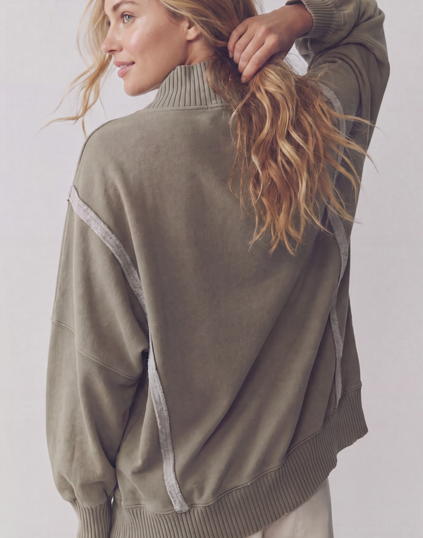 A woman wearing an oversized ribbed knit sweater in a muted olive green color, shown from the back with her hair pulled back. The sweater features contrasting light gray ribbed trim along the shoulders and sides.