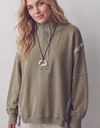 A woman wearing a sage green mock-neck sweatshirt with ribbed cuffs and hem, featuring a decorative geometric embroidery on the chest and a large statement ring necklace.
