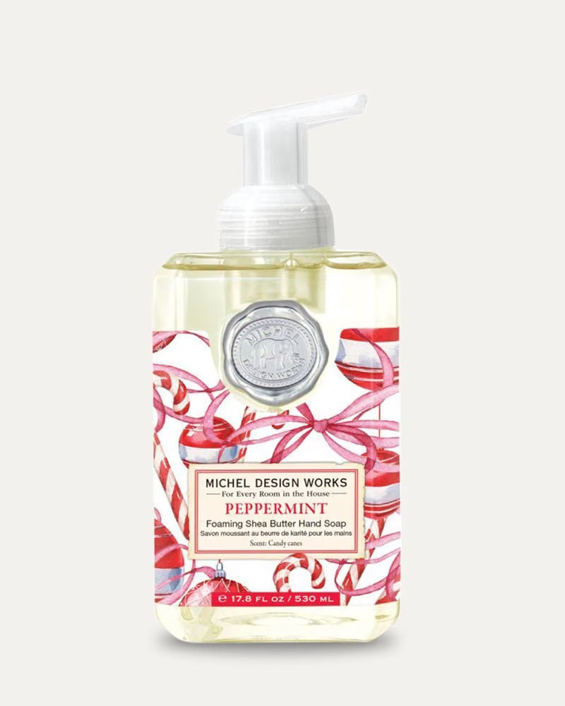 Michel Design Works Peppermint Foaming Shea Butter Hand Soap in a clear bottle with a white pump dispenser, decorated with red and white candy cane illustrations on a light beige background.