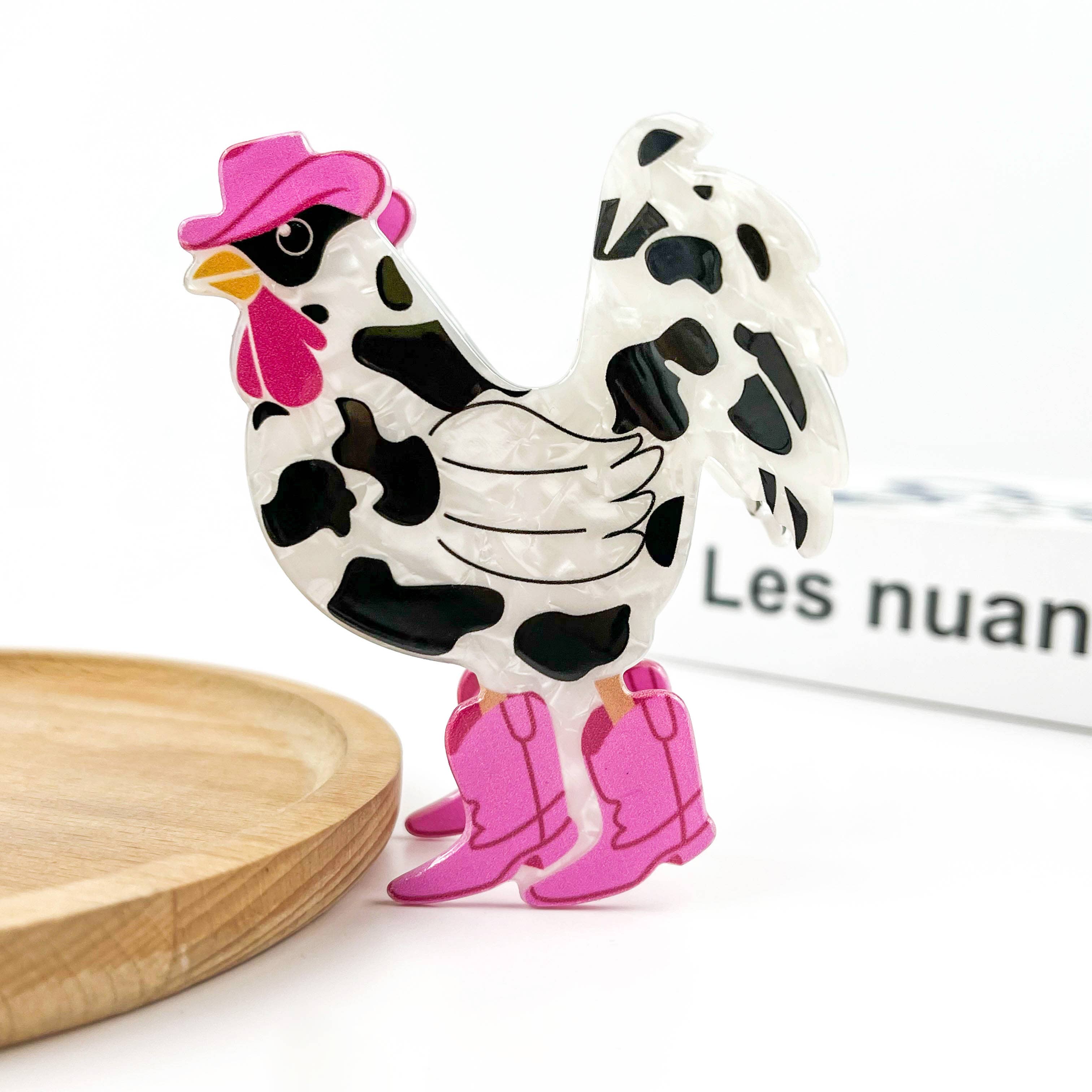 A stylized rooster clip with a white body, black cow-print spots, a pink hat, and pink boots, displayed on a white surface with a wooden plate edge and a blurred book in the background.