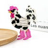 A stylized rooster clip with a white body, black cow-print spots, a pink hat, and pink boots, displayed on a white surface with a wooden plate edge and a blurred book in the background.