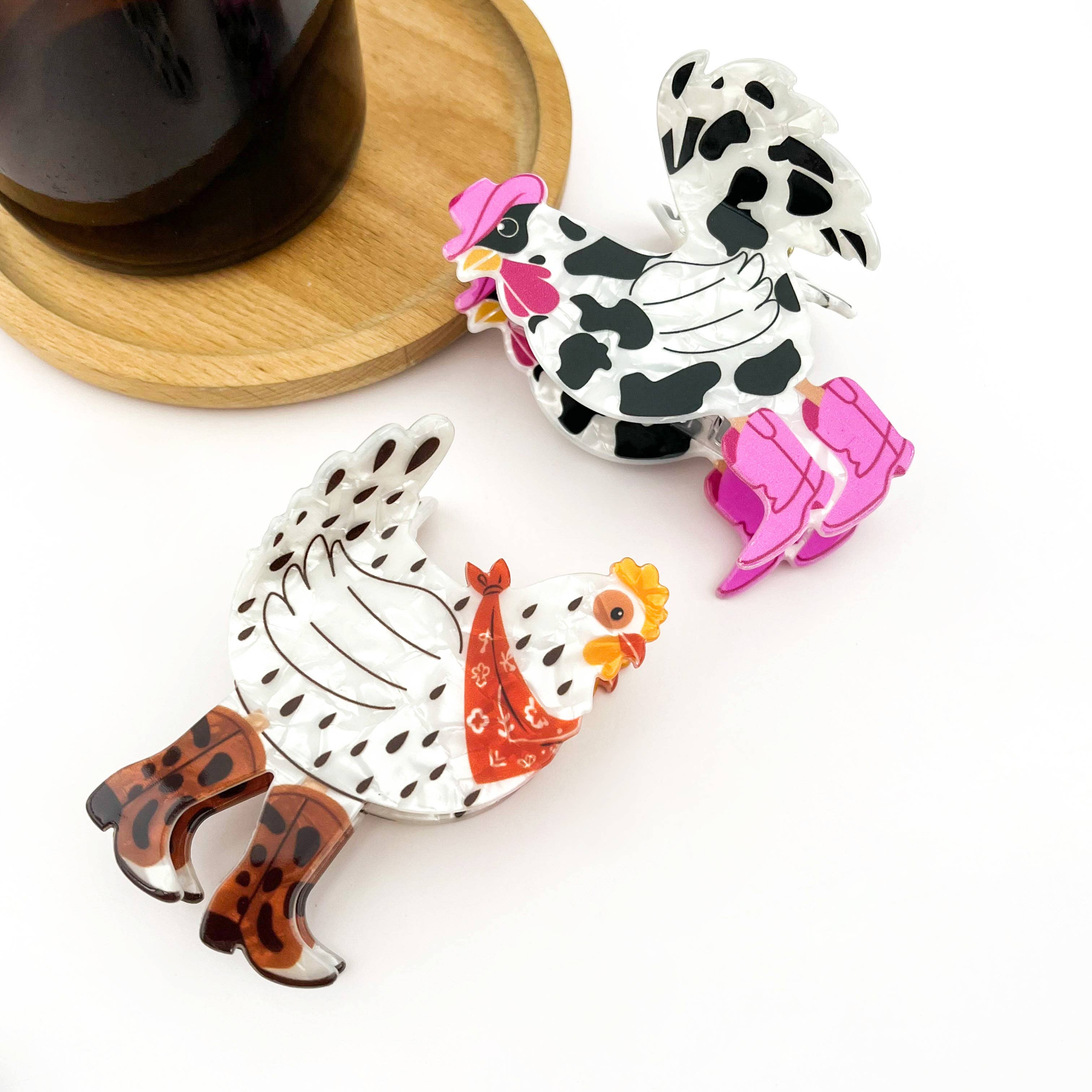 Two cartoon-style chicken-shaped claw clips on a white background, with one black-and-white spotted chicken wearing pink boots and the other white with black spots, an orange comb, a red bandana, and brown leopard-print boots. A dark cup on a wooden saucer is partially visible in the upper left.
