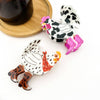 Two cartoon-style chicken-shaped claw clips on a white background, with one black-and-white spotted chicken wearing pink boots and the other white with black spots, an orange comb, a red bandana, and brown leopard-print boots. A dark cup on a wooden saucer is partially visible in the upper left.