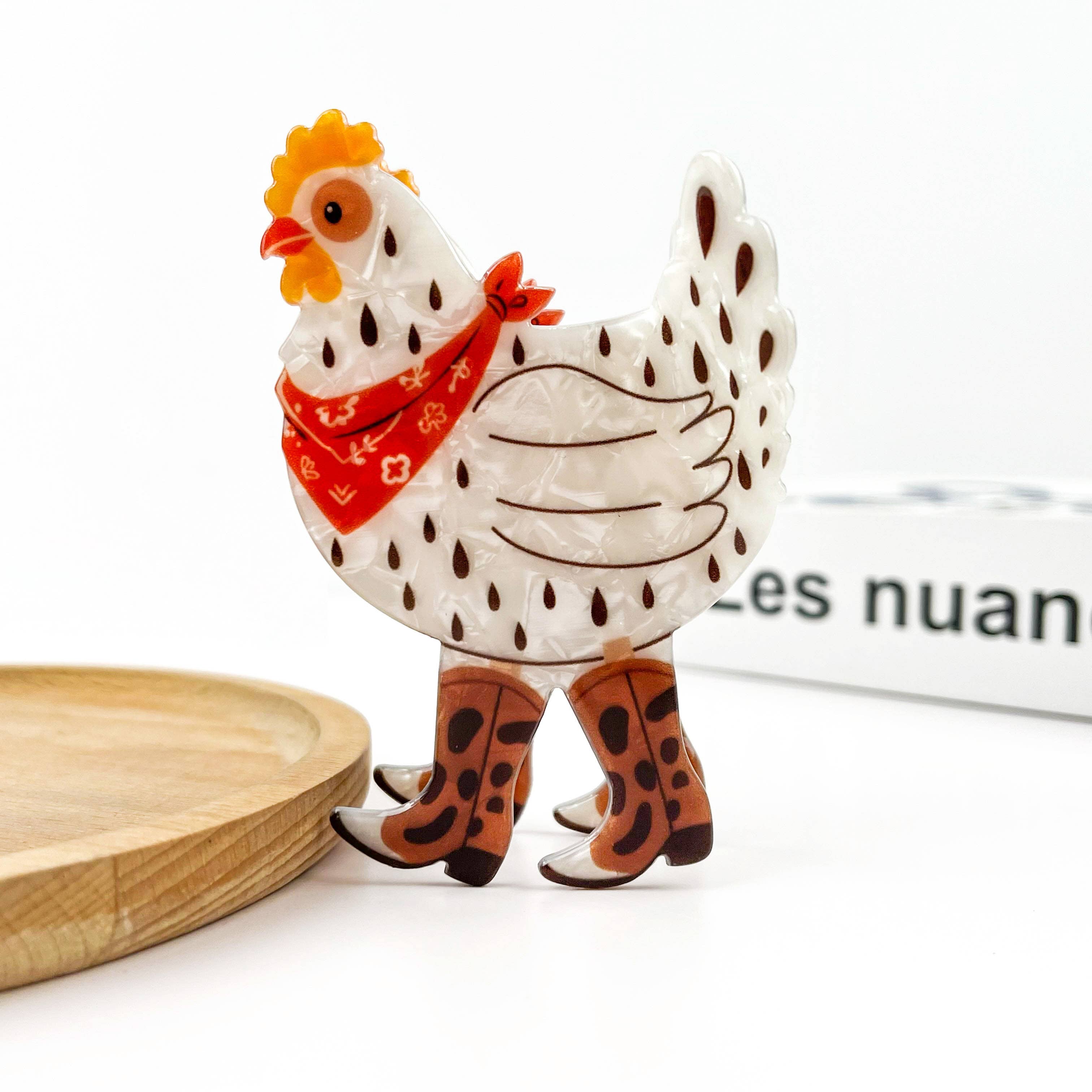 A decorative chicken clip with a white body, brown spots, a red bandana, and brown cowboy boots, standing on a white surface with a wooden tray edge visible.