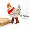 A decorative chicken clip with a white body, brown spots, a red bandana, and brown cowboy boots, standing on a white surface with a wooden tray edge visible.