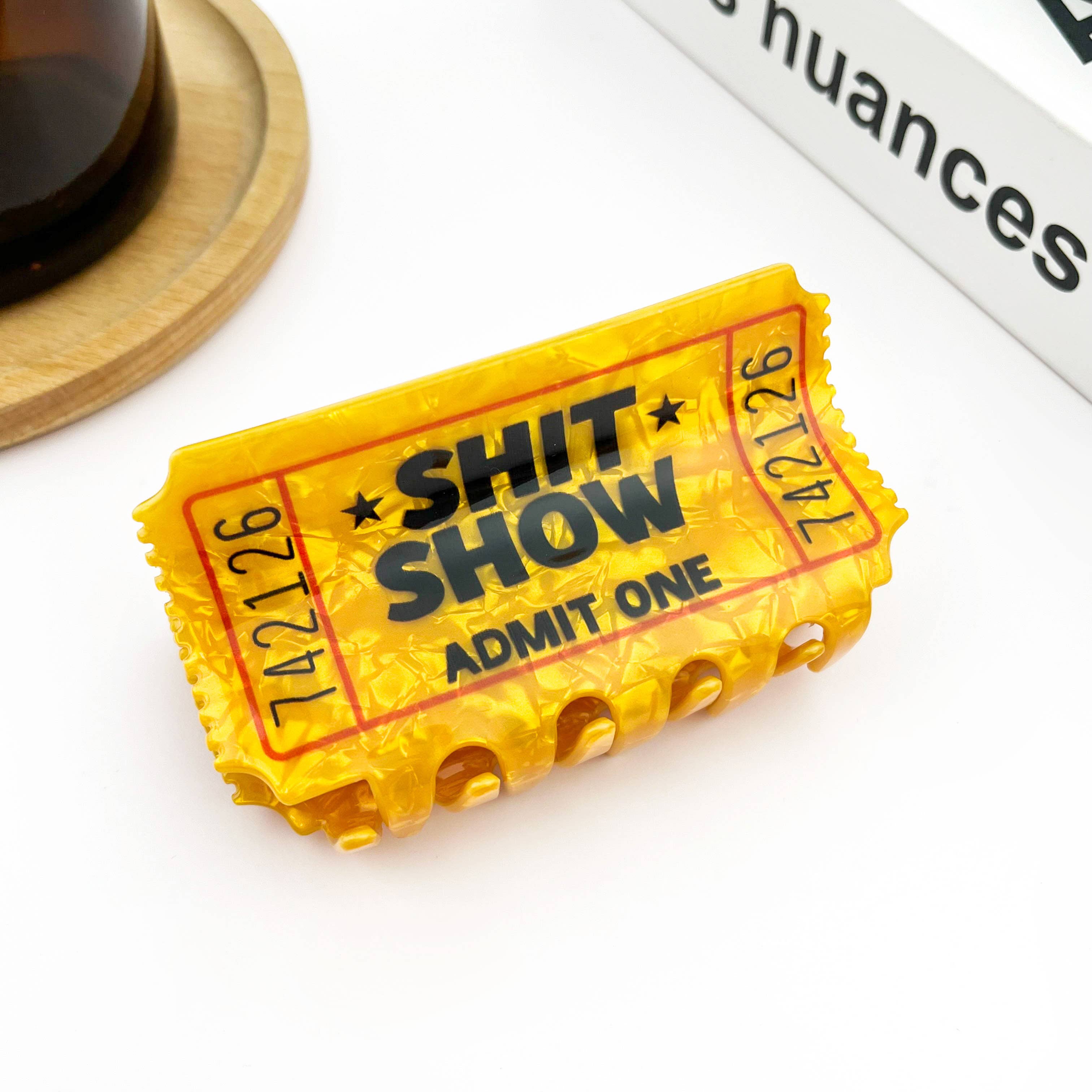 A novelty yellow plastic 'Admit One' ticket holder for a 'Shit Show' event, styled hair clip on a white surface with a wooden coaster and a book in the background.