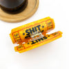 A novelty hair clip designed to look like a yellow and red carnival ticket with bold black text reading 'SHIT' and numbers, shown on a white background with a wooden coaster and dark glass partially visible in the background.