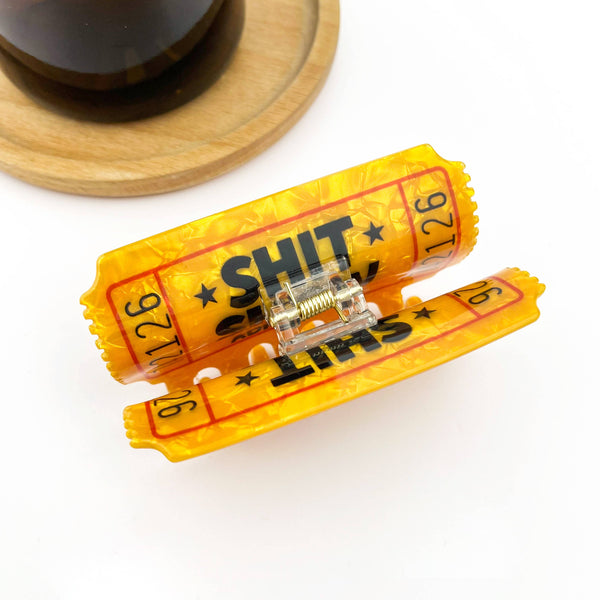 A novelty hair clip designed to look like a yellow and red carnival ticket with bold black text reading 'SHIT' and numbers, shown on a white background with a wooden coaster and dark glass partially visible in the background.