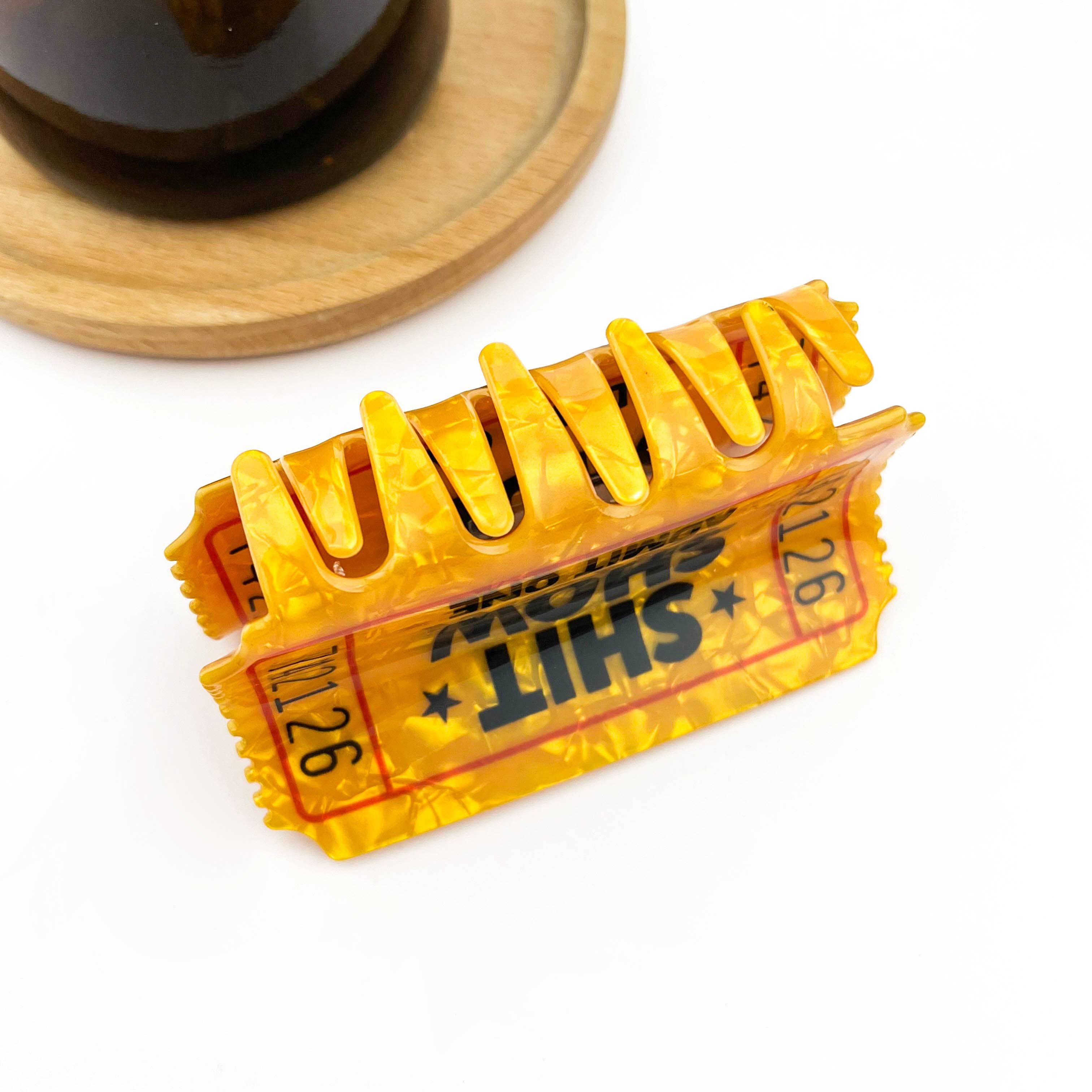 A translucent yellow-orange hair claw clip styled like a vintage movie ticket with bold black text reading 'SHT SHOW' and red border details, photographed on a white background with a wooden coaster and dark bottle partially visible in the background.