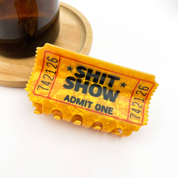 A novelty yellow and orange plastic 'Shit Show Admit One' ticket claw clip, styled on a white surface with a wooden coaster and a dark glass bottle in the background.