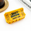 A novelty yellow plastic 'Admit One' ticket holder for a 'Shit Show' event, styled hair clip on a white surface with a wooden coaster and a book in the background.