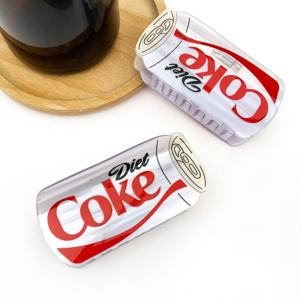 Two Diet Coke claw clip cans on a white surface with a wooden coaster and glass in the background.